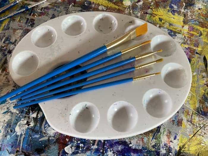 A set of blue handled acrylic paint brushes for beginners resting on top of a white palette with 10 wells. 