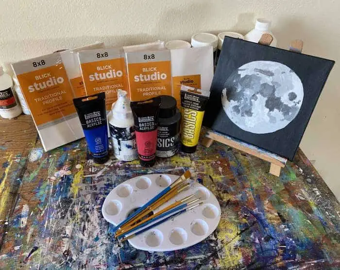 Three eight by eight inch blick studio canvases fanned out in the background. A row of Liquitex paints of primary colors blue, red, and yellow as well as black and white. A 10 well palette with 6 paint brushes on it and an easel with a painting of the moon displayed. 