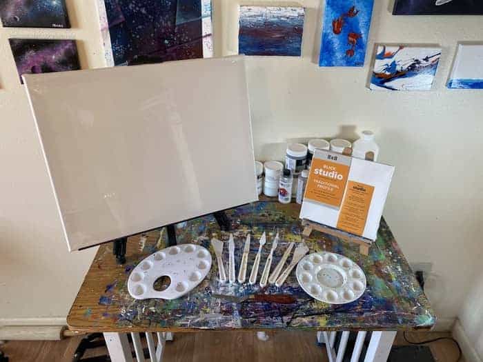 A large and small easel each holding a canvas behind a set of palette knives fanned out. There are also two different types of palettes on either side of the fanned out palette knives. 