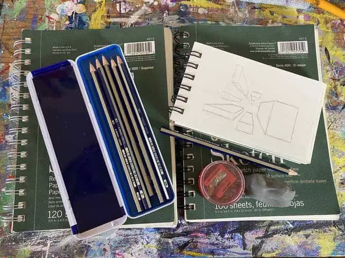 Two sketch pads laid side by side. On top of the sketch pads are a pencil set, a clay eraser, a pencil sharpener, and a smaller sketch pad that has a sketch on perspective displayed. 