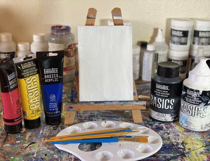 A blank canvas waiting to be painting. Flanked on either side by the basic colors needed to make any color. Palette and paintbrushes laid at the feet of the easel holding the canvas. 